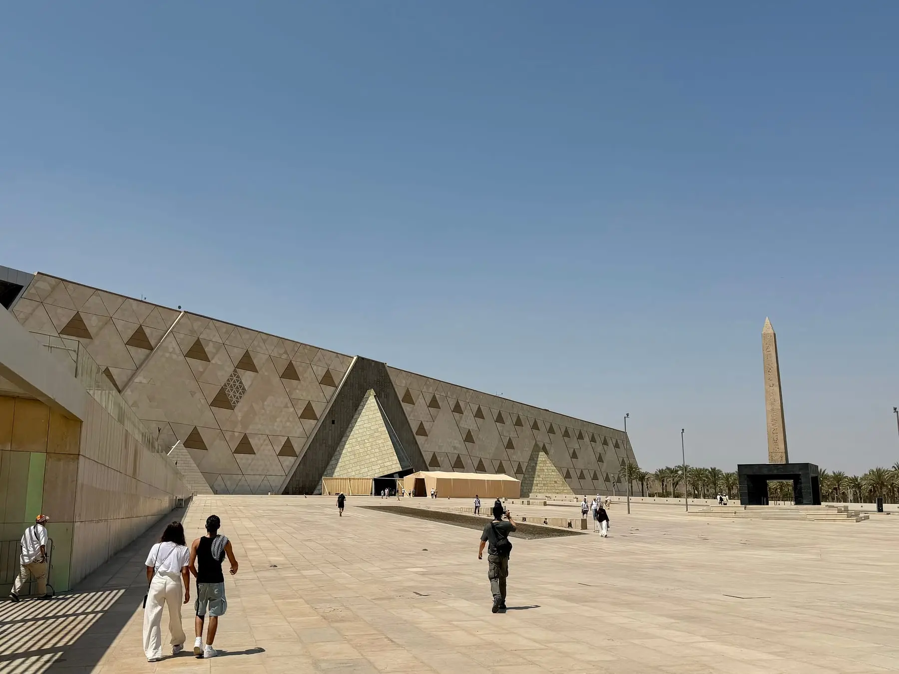 The Grand Egyptian Museum's triangular stone facade and obelisk in Giza, with visitors crossing the plaza